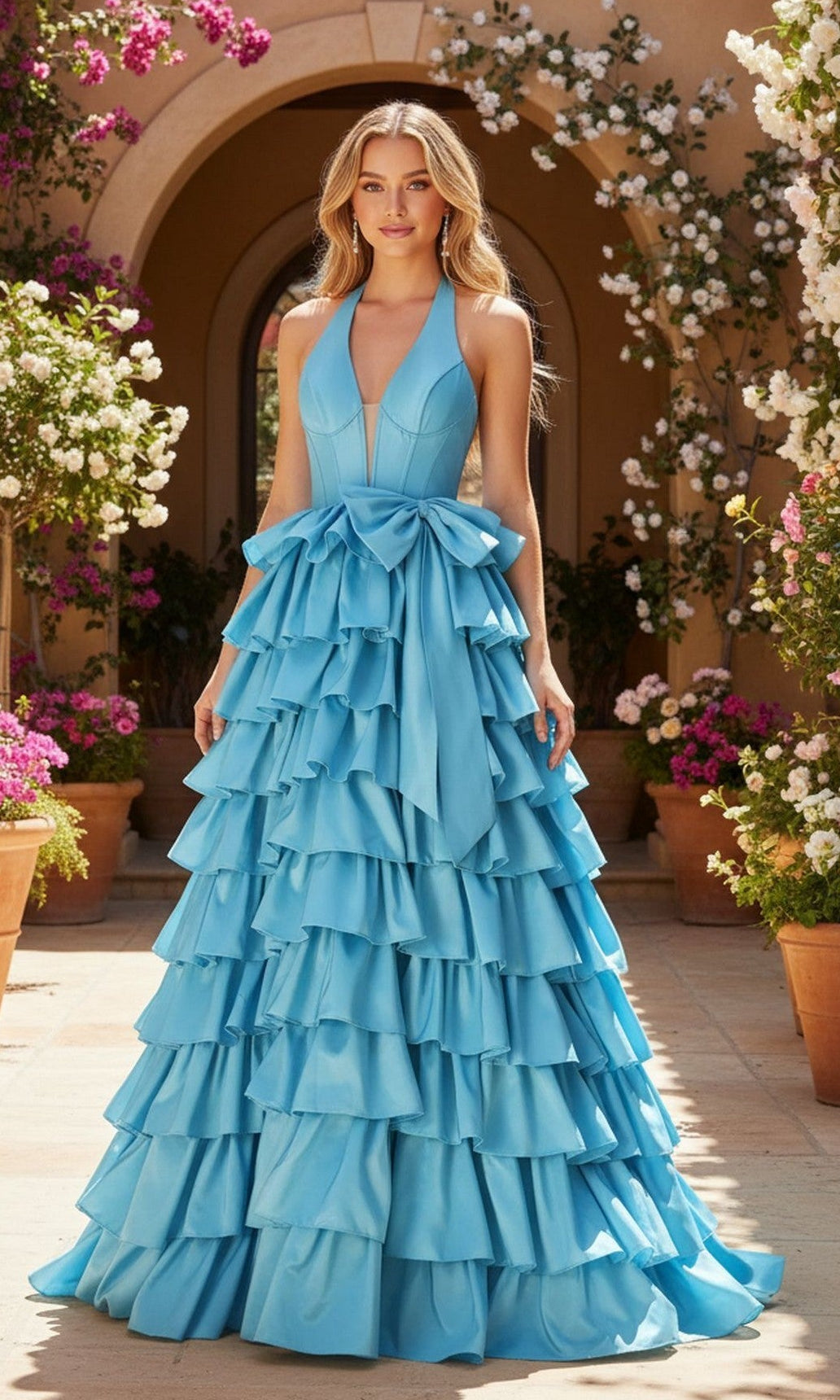 A woman stands outdoors in a light blue Halter Prom Ball Gown (PromGirl Exclusive 250045), surrounded by flowering plants in terracotta pots.
